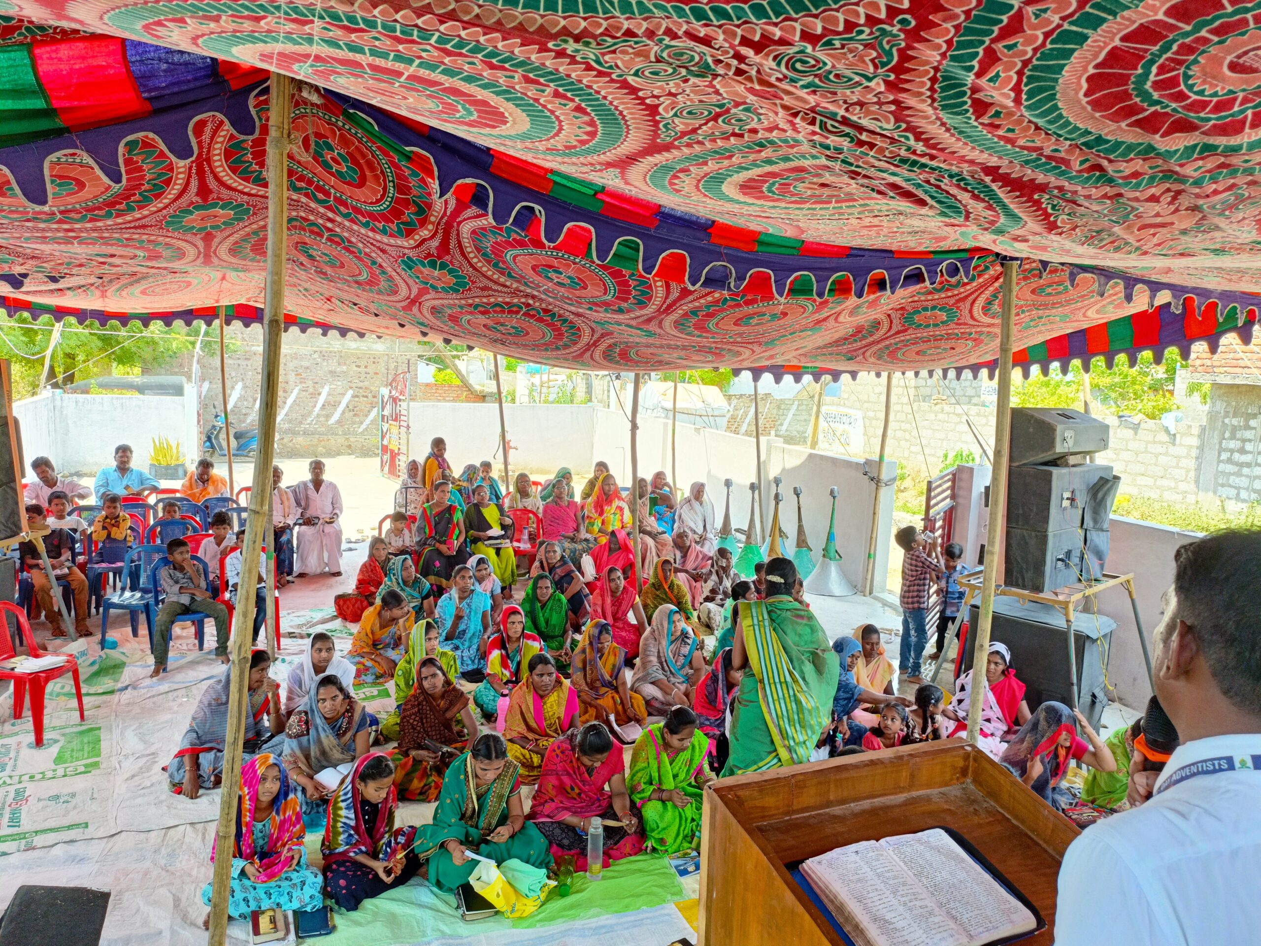 A village gathering under a decorated canopy, people seated on mats before a speaker at a wooden pulpit