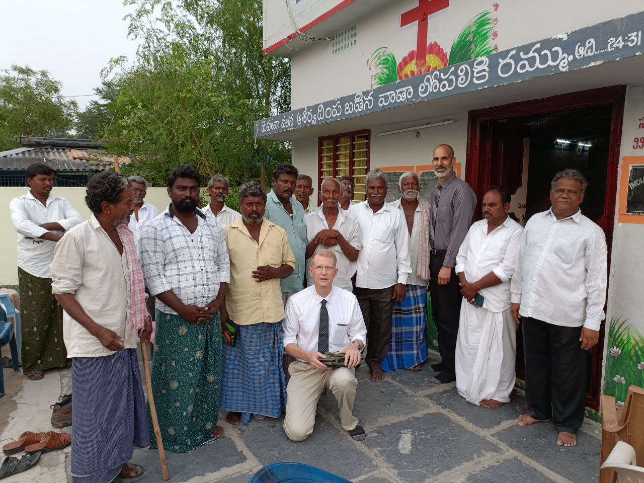 A visiting pastor kneeling with local believers outside a village church